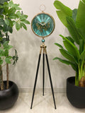 Floor clock with a turquoise face and gold accents on a tripod stand, surrounded by potted plants.