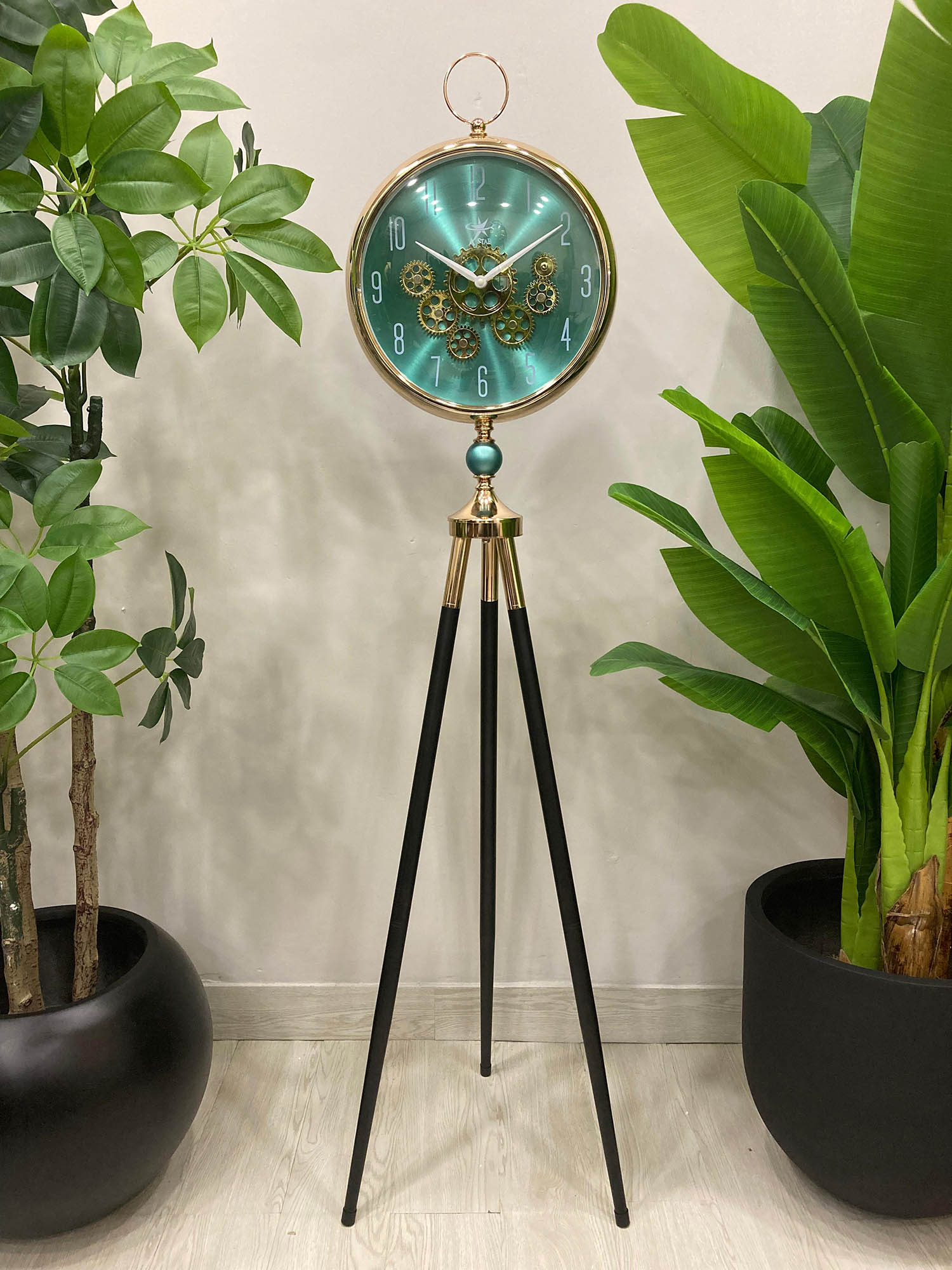 Floor clock with a turquoise face and gold accents on a tripod stand, surrounded by potted plants.