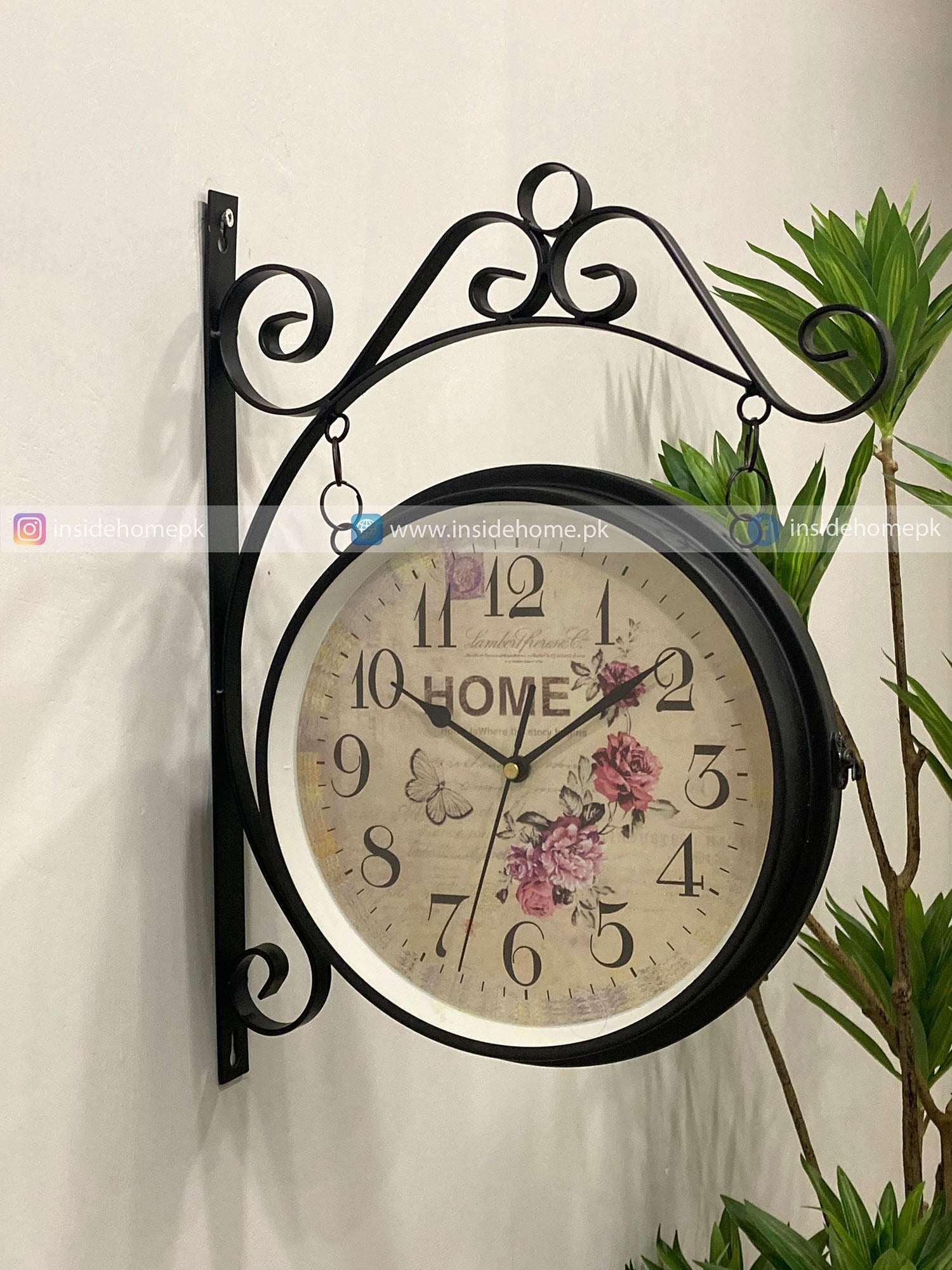 Decorative wall clock with floral design and 'Home' text on a white wall with a plant.