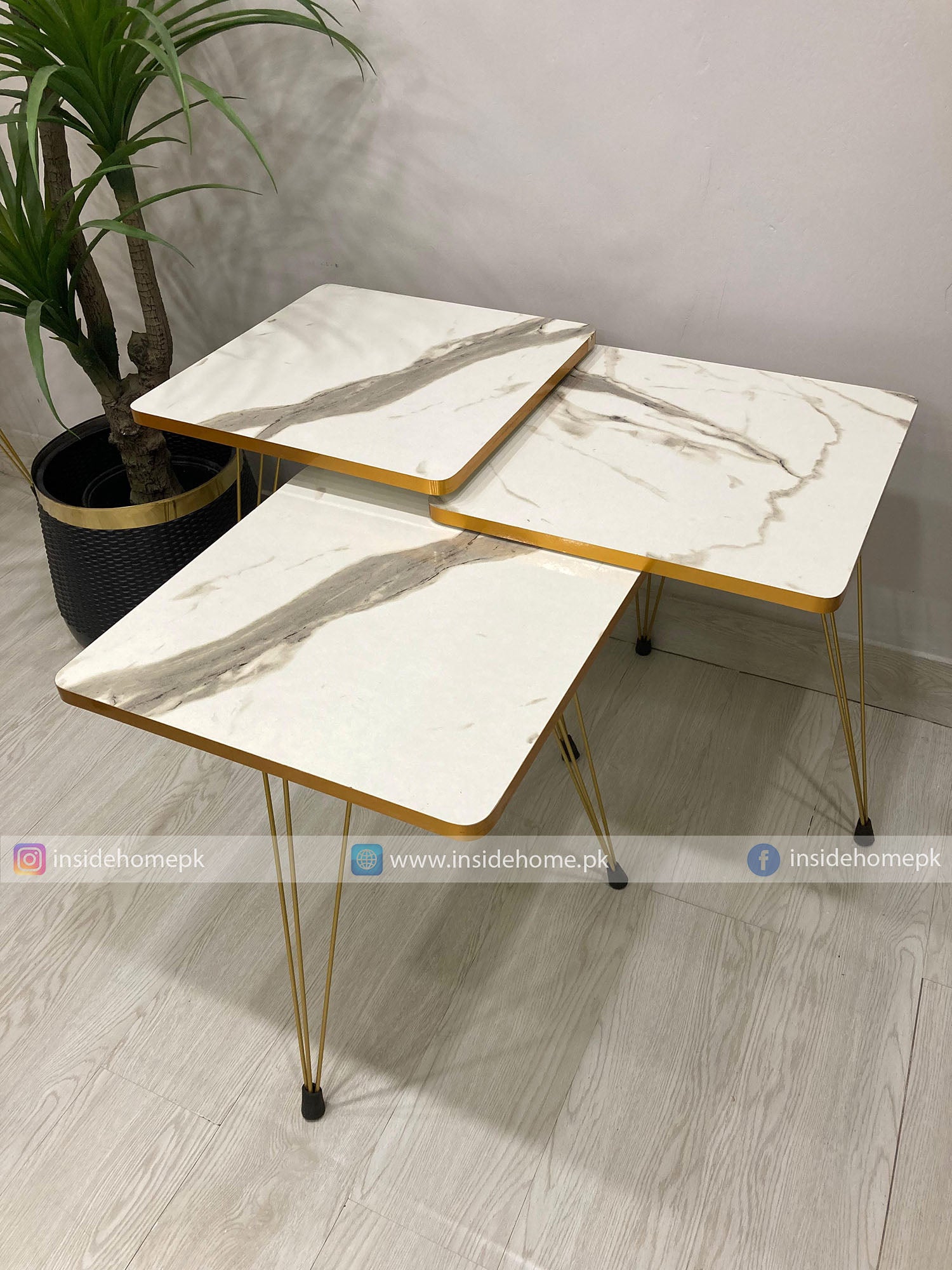 Two marble-top tables with gold legs on a wooden floor, with a plant in the background.