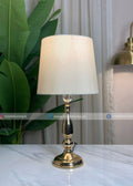Table lamp with gold base and white shade on a marble surface, next to a plant and curtain.