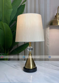 Table lamp with a gold base and white shade on a marble surface, with plants and a curtain in the background.