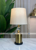 Table lamp with a white shade and gold base on a marble surface, with a plant and curtain in the background.