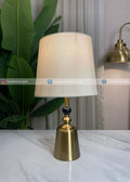Table lamp with beige shade and brass base on a marble surface, with a plant and curtain in the background.