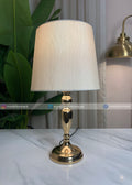 Table lamp with gold base and white shade on a marble surface, with plants and another lamp in the background.